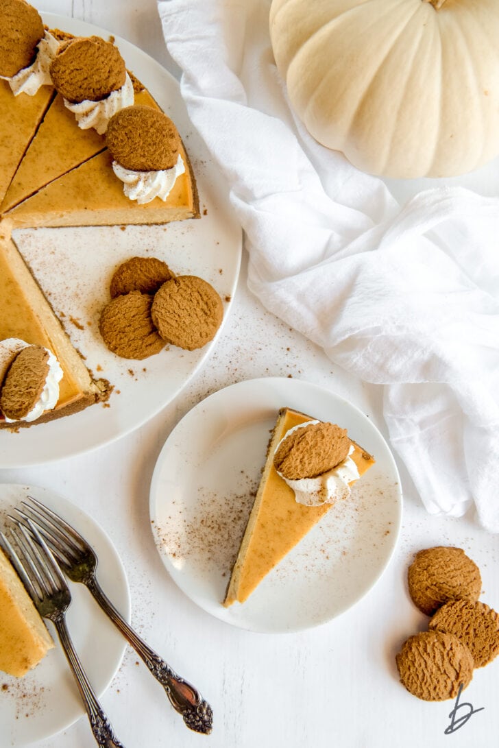 slice of pumpkin cheesecake on white plate next to the full cheesecake garnished with gingersnap cookies