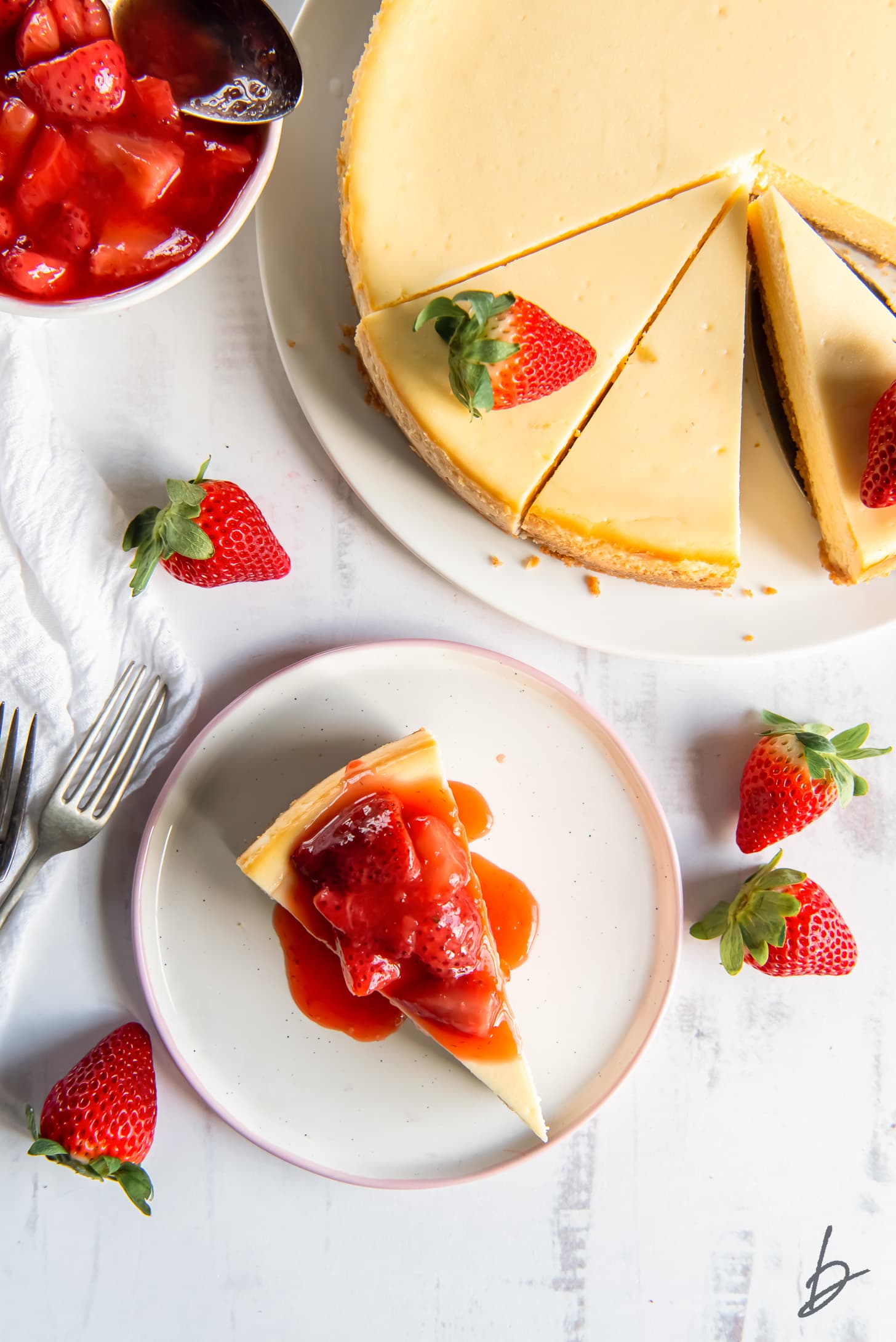 slice of cheesecake topped with strawberry sauce on plate next to whole cheesecake and fresh strawberries