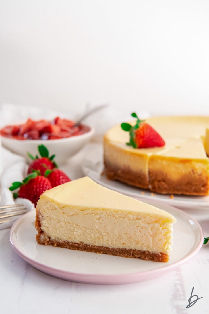 slice of plain cheesecake on plate in front of whole cheesecake and fresh strawberries