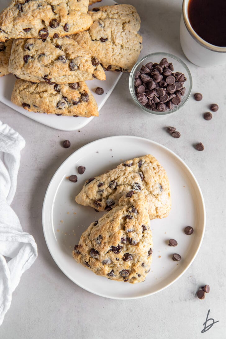 two chocolate chip scones on a white plate next to more scones on another plate and small bowl of chocolate chips