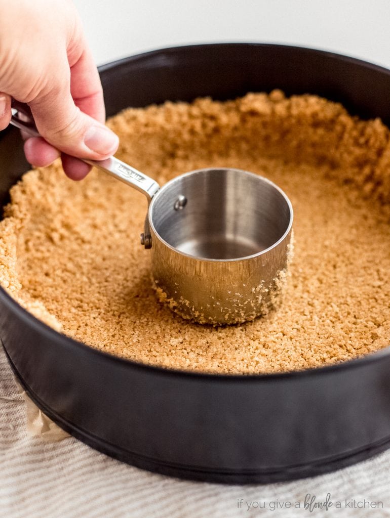 hand pressing stainless steel measuring cup on graham cracker crust in springform pan