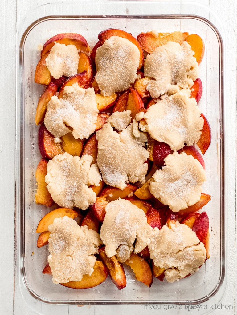 cinnamon sugar sprinkled on top of unbaked cobbler dough on top of peaches in glass baking pan