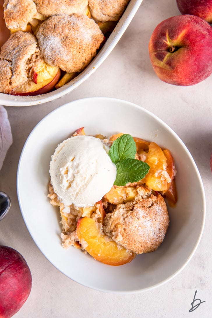peach cobbler serving in a bowl with scoop of vanilla ice cream next to fresh peaches