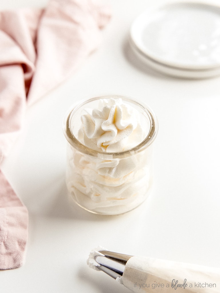 homemade whipped cream in a glass jar. piping bag and pink kitchen towel on white surface