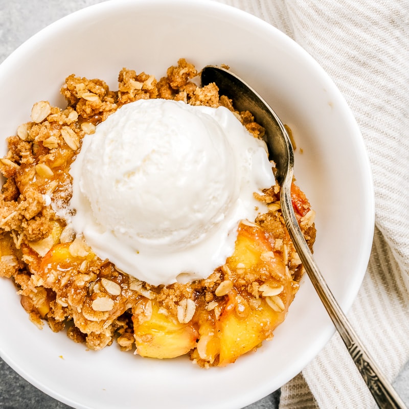 bowl of peach crisp with scoop oof vanilla ice cream in white bowl with spoon