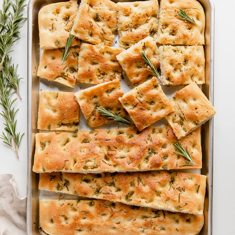 focaccia bread cut into squares on baking sheet with rosemary sprigs