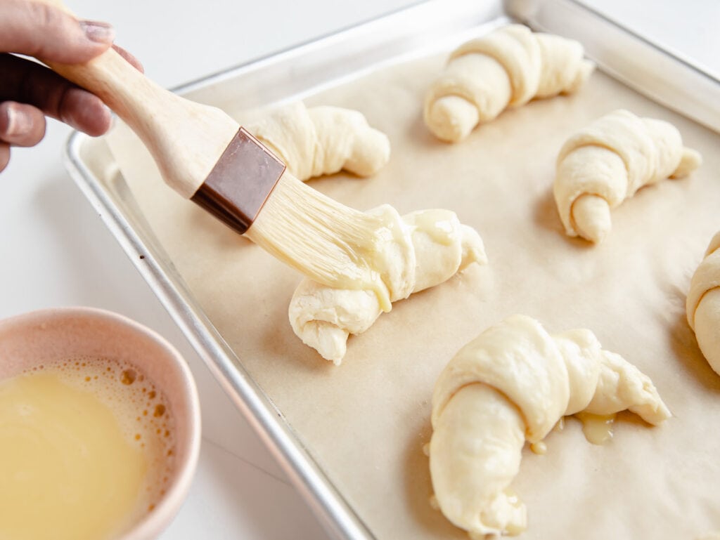 pastry brush adding egg wash to unbaked croissants on baking sheet