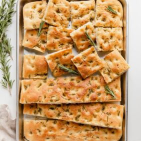 focaccia bread on large baking pan; half the bread cut into squares; rosemary next to pan