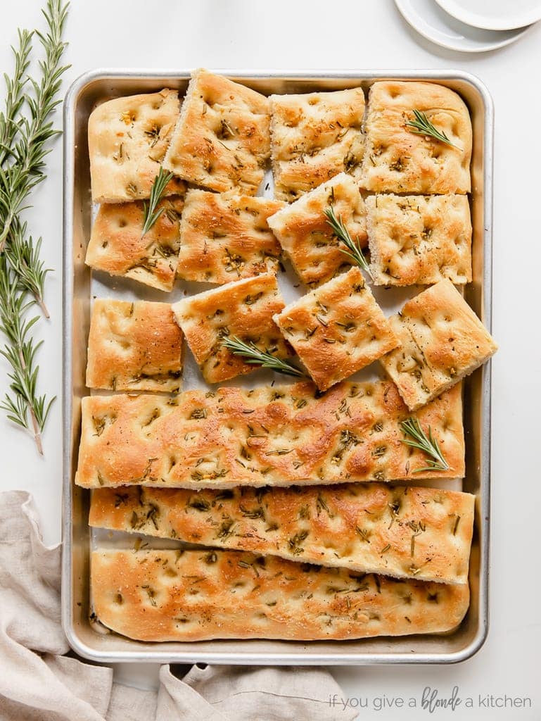 focaccia bread on large baking pan; half the bread cut into squares; rosemary next to pan