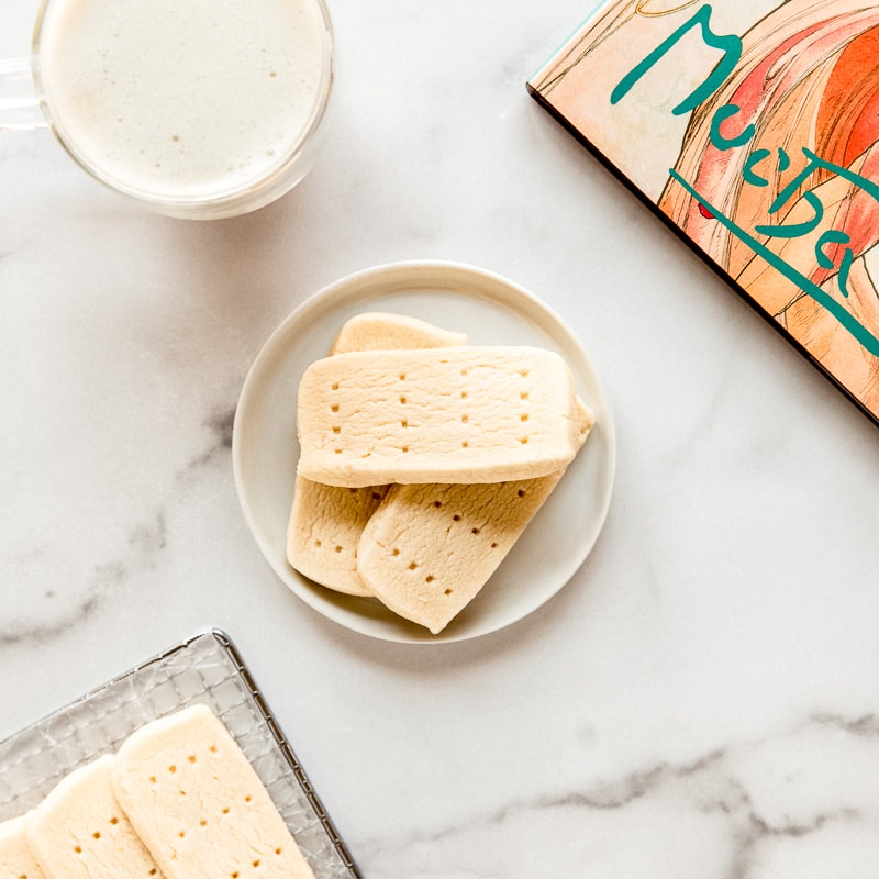 three shortbread cookies on round white plate, cup of coffee and book on marble countertop