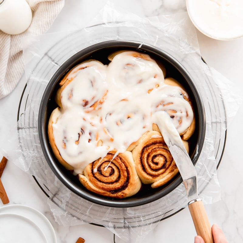 offset spatula spreading cream cheese icing on cinnamon rolls in round cake pan
