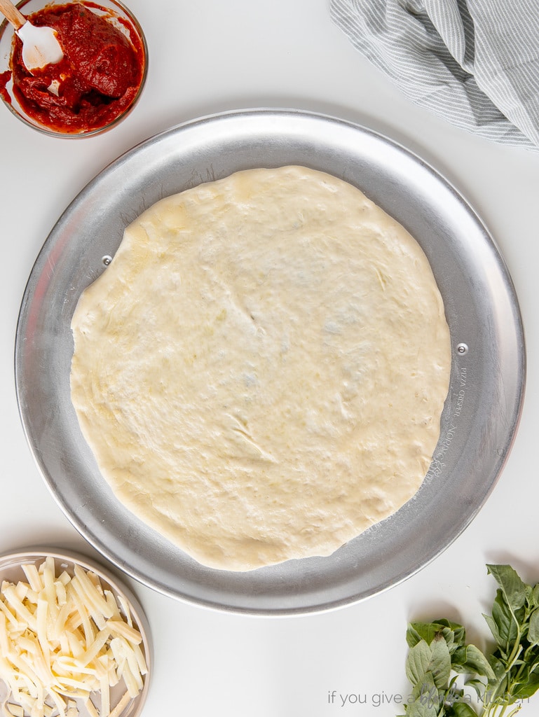 pizza dough stretched out on metal pizza pan. Bowls of sauce, cheese and basil next to pan