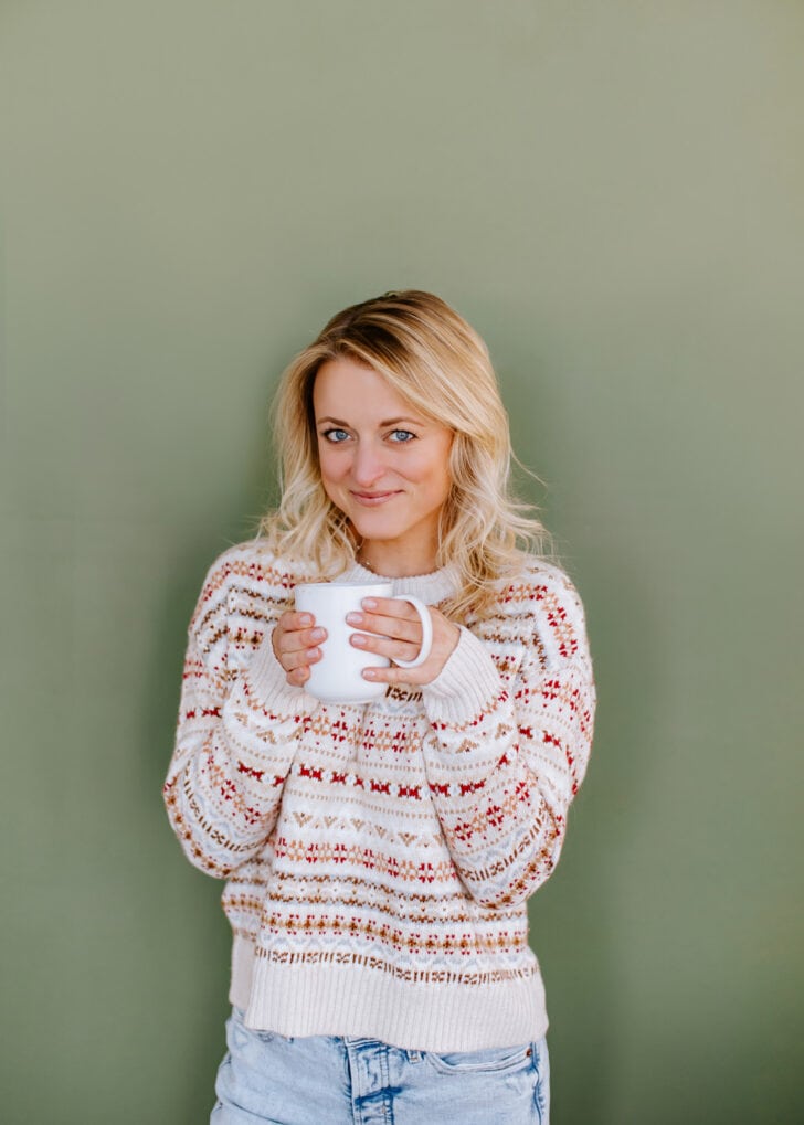green wall with blonde woman in a white patterned holiday sweater holding a mug with both hands