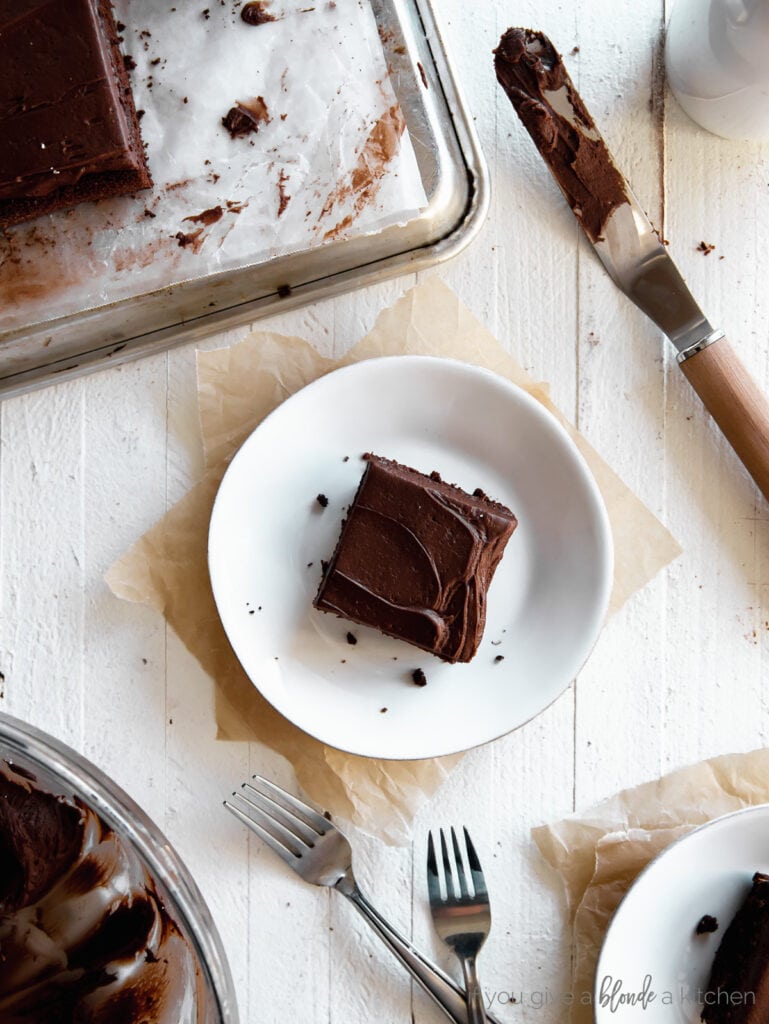 slice of chocolate sheet cake with chocolate frosting on a round plate next to forks and offset spatula