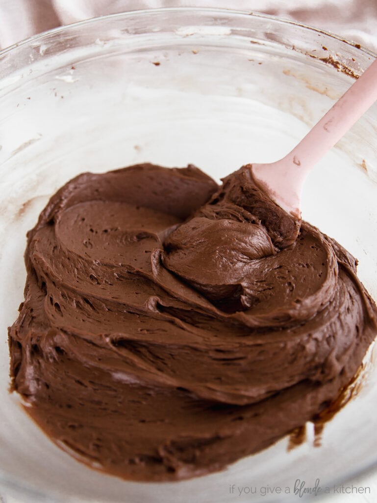 glass bowl filled with chocolate butter cream frosting and a pink rubber spatula