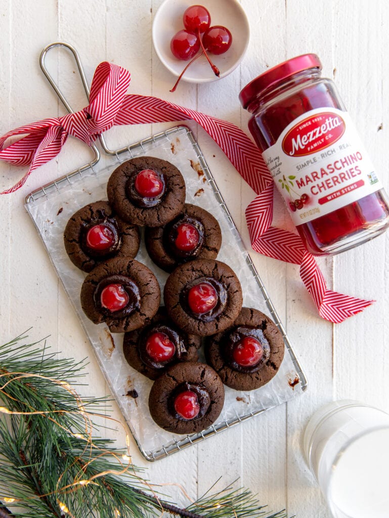 chocolate cherry thumbprint cookies on cooling rack with red holiday ribbon; jar of cherries next to cooling rack
