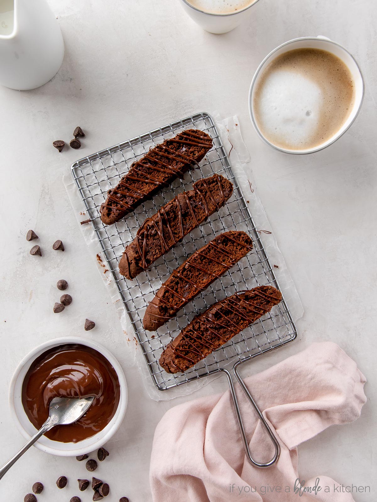 four chocolate biscotti on a small wire cooling rack next to coffee and bowl of melted chocolate