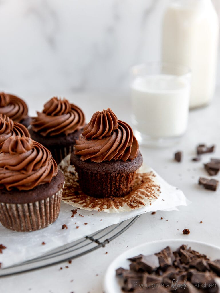 chocolate cupcake with chocolate frosting on open paper cupcake liner on wire cooling rack with more cupcakes