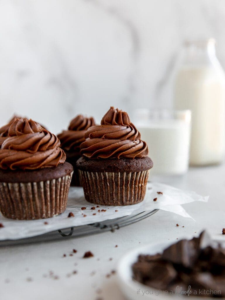 chocolate cupcake with chocolate frosting on wire rack with glass bottle of milk behind cupcakes