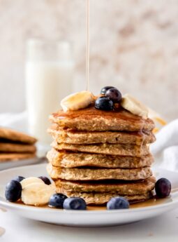maple syrup poured on a stack of banana oatmeal pancakes with sliced bananas and blueberries