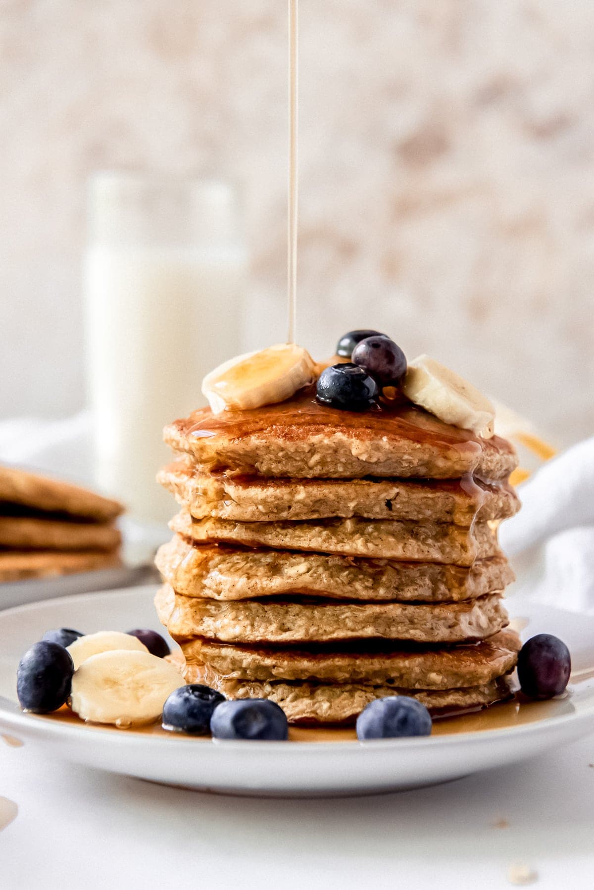 maple syrup poured on a stack of banana oatmeal pancakes with sliced bananas and blueberries