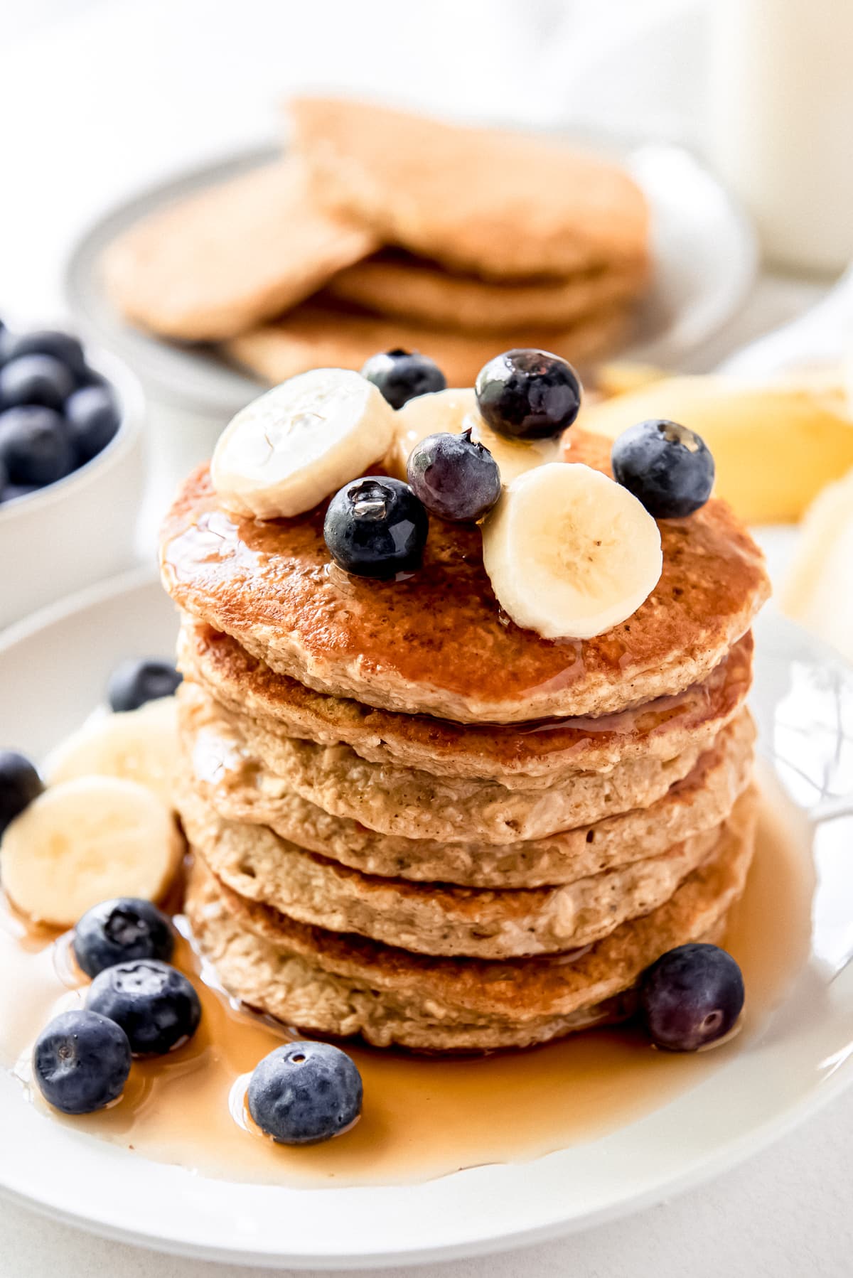 stack of banana oatmeal pancakes with sliced bananas, blueberries and syrup on top.