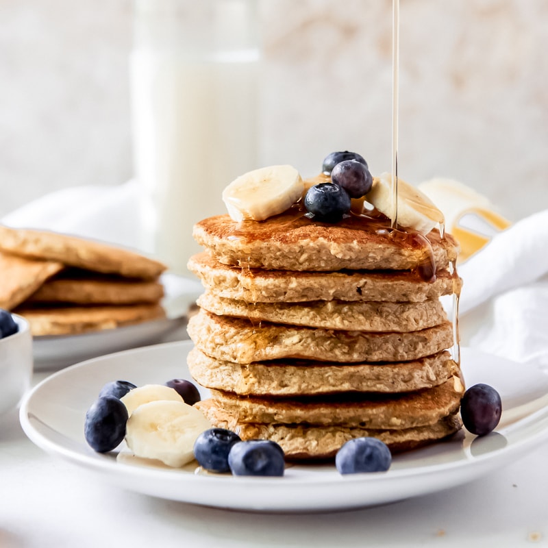 stack of banana oatmeal pancakes with blueberries and banana slices on top, syrup dripping down