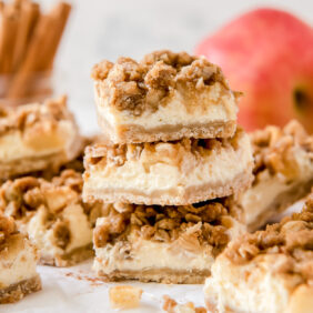 stack of three apple cheesecake bars in front of more bars and apples