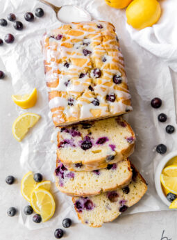 lemon blueberry bread with three slices cut off the end on top of parchment paper with fresh blueberries and lemon slices