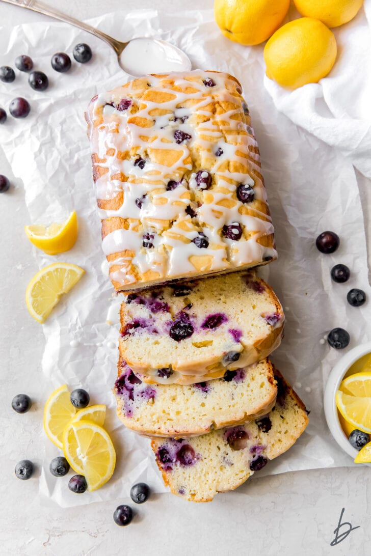 lemon blueberry bread with three slices cut off the end on top of parchment paper with fresh blueberries and lemon slices