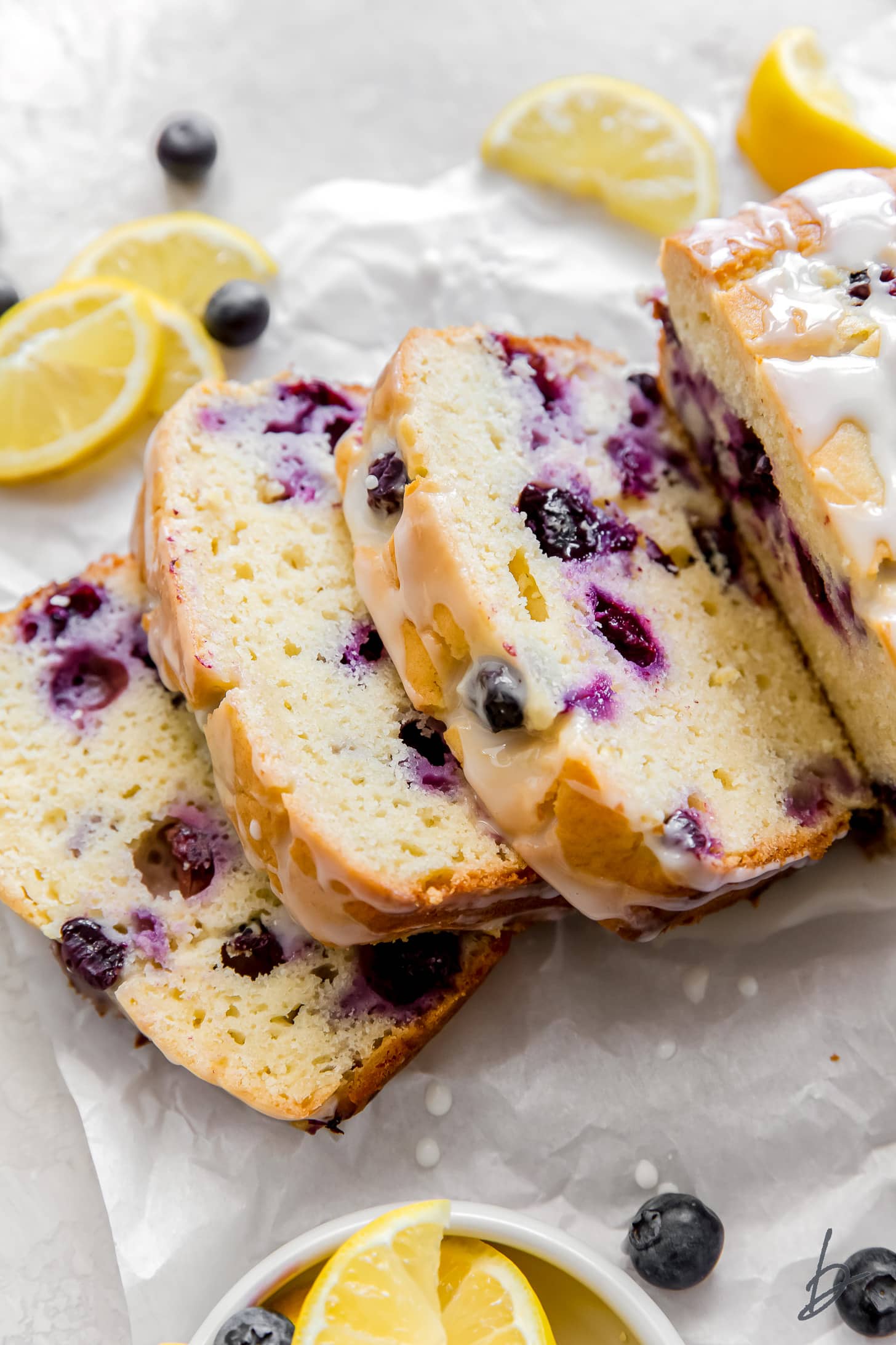Three slices of lemon blueberry bread cut off a loaf on top of parchment paper with lemon slices