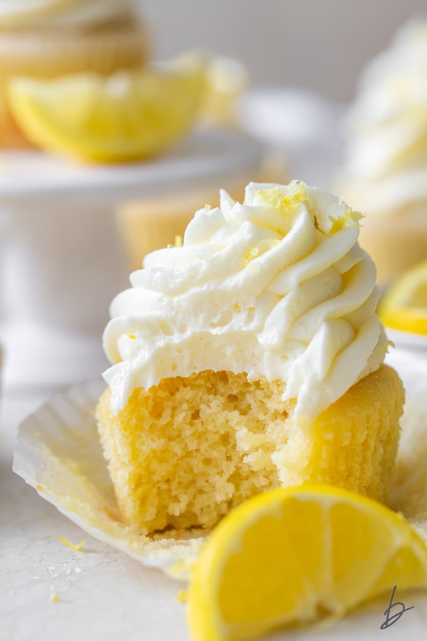 bite taken out of lemon cupcake sitting on top of paper cupcake liner with lemon slice in front.