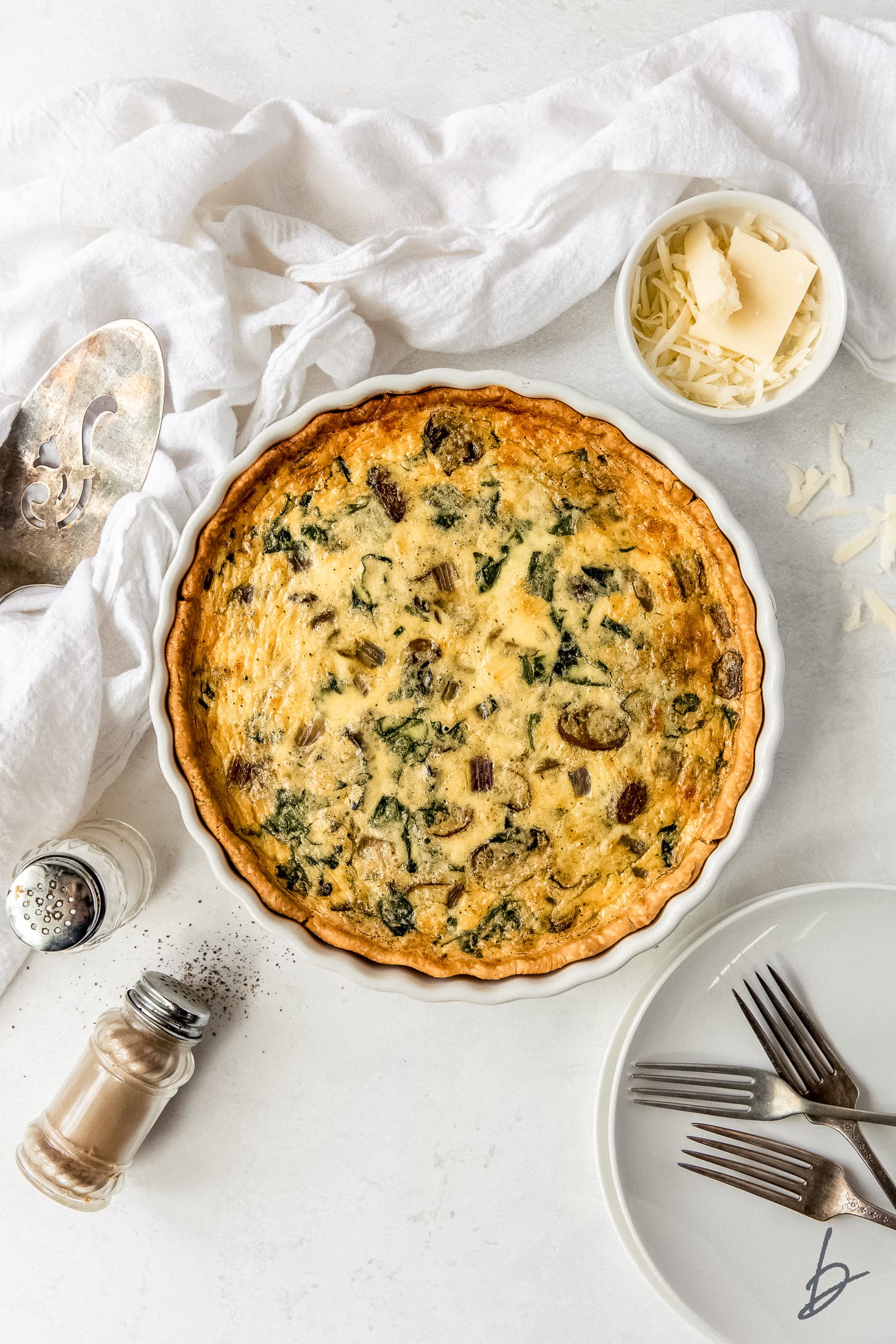 spinach mushroom quiche in a white ceramic quiche next to plates, forks and salt and pepper shakers