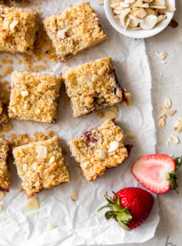 strawberry oatmeal bars on parchment paper next to fresh strawberries and small bowl of slivered almonds