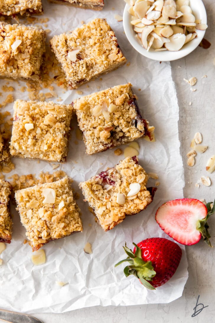 strawberry oatmeal bars on parchment paper next to fresh strawberries and small bowl of slivered almonds