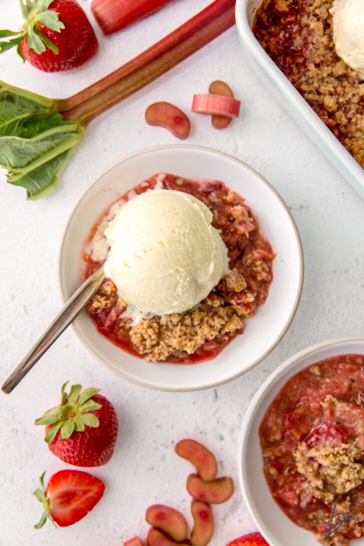 strawberry rhubarb crisp in a bowl with a scoop of vanilla ice cream and spoon next to strawberries