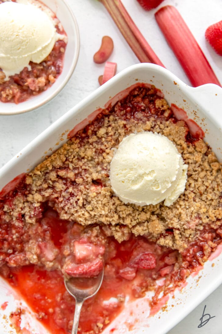 strawberry rhubarb crisp in a white baking dish with a spoon scooping crisp and a scoop of vanilla ice cream