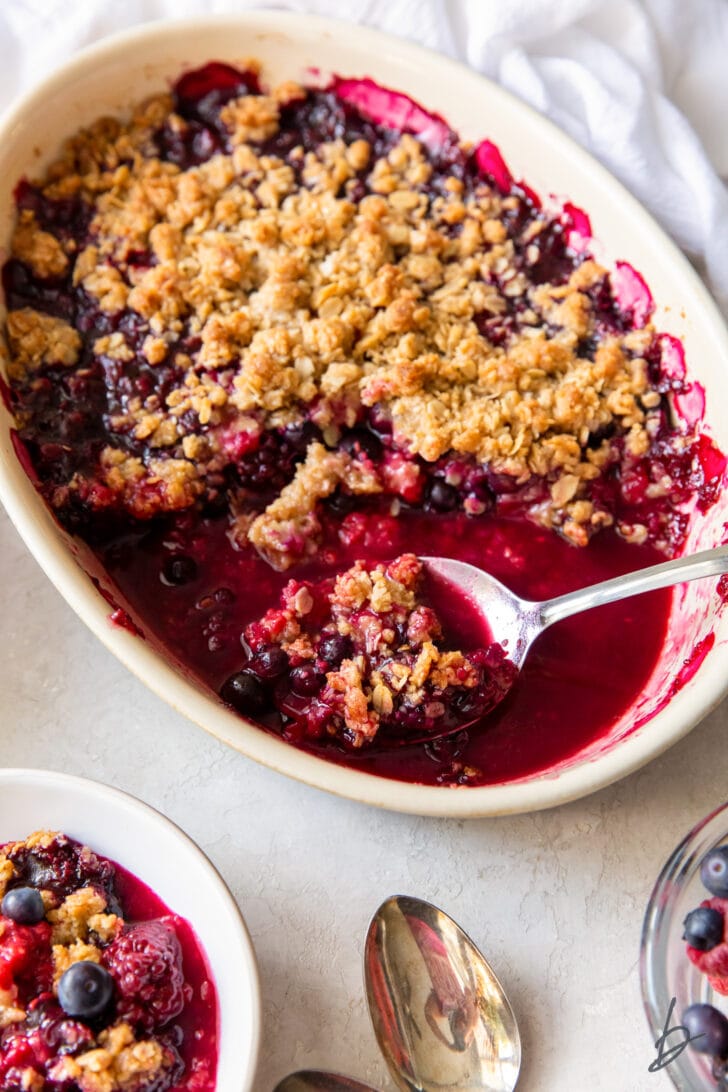 triple berry crisp in an oval baking dish with serving spoon holding some crisp