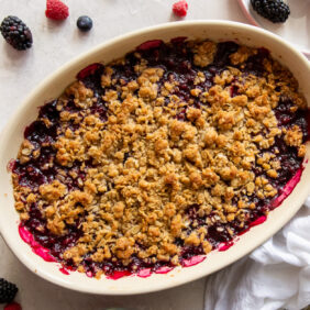 triple berry crisp in an oval baking dish next to white linen cloth and fresh berries