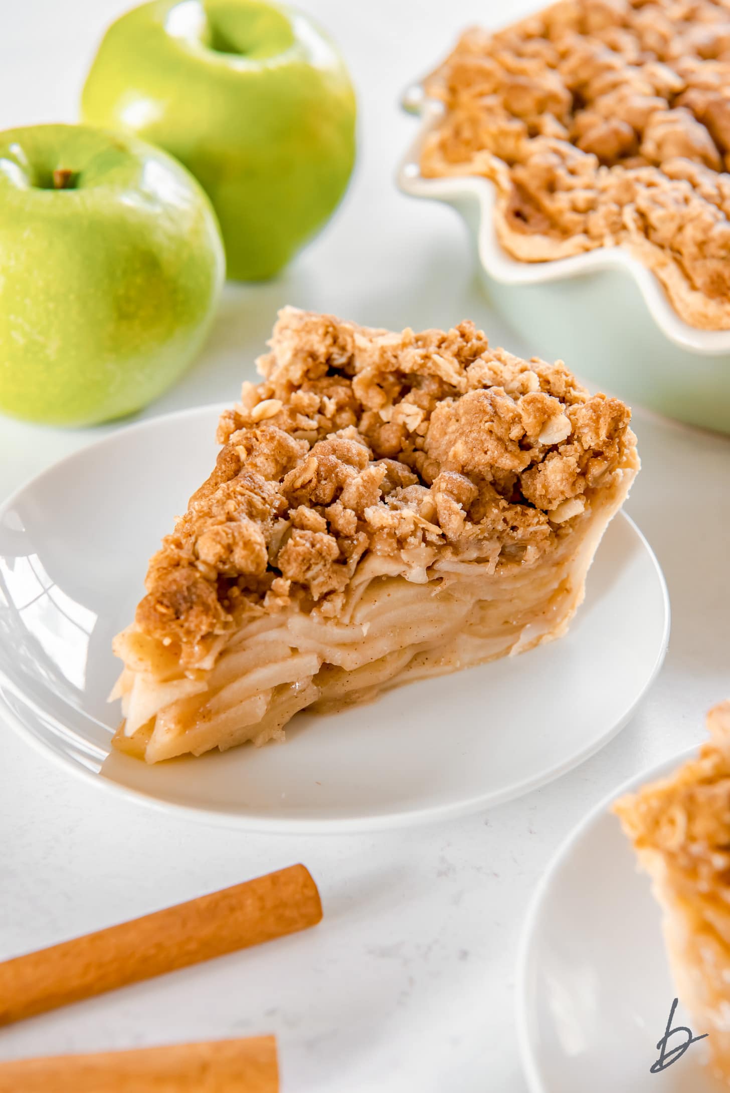 slice of apple crumble pie on white round plate in front of granny smith apples and cinnamon sticks