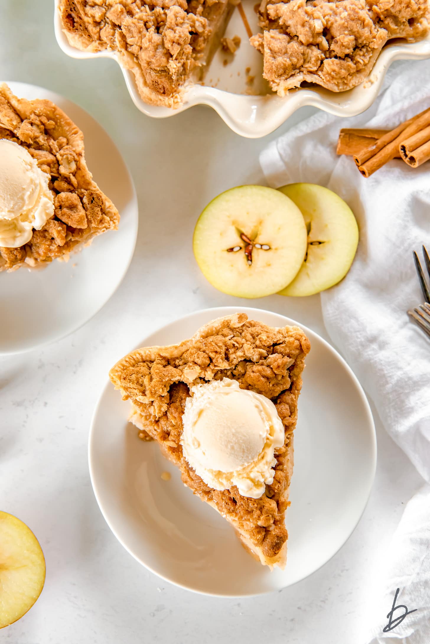 apple crumble pie slice topped with scoop of vanilla ice cream on white round plate next to apple slices and cinnamon sticks