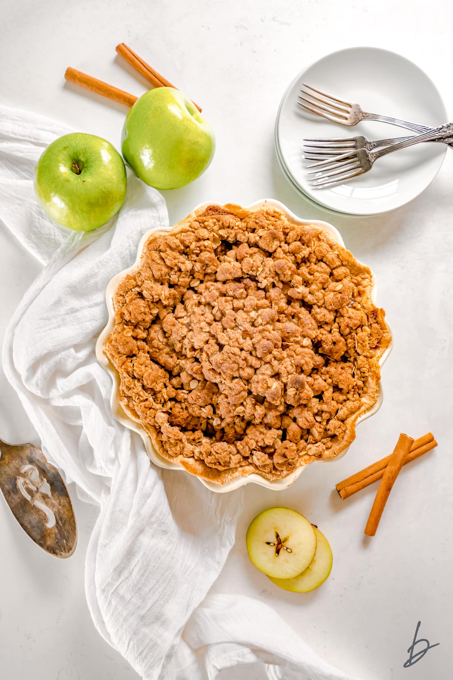 apple crumble pie in a pie dish next to white kitchen cloth, granny smith apples and cinnamon sticks