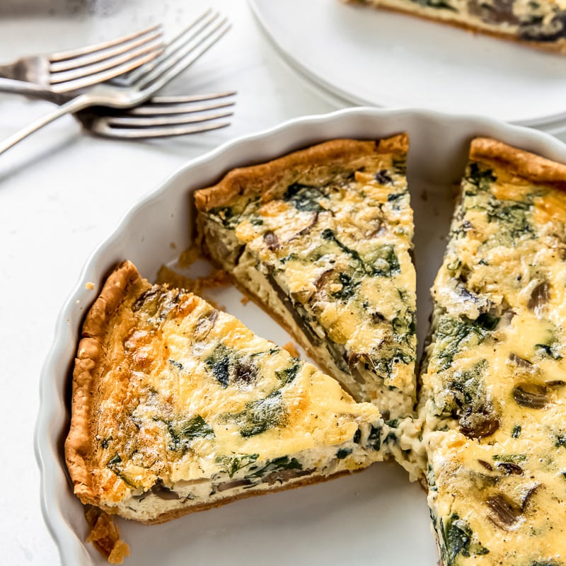 spinach mushroom quiche in a white ceramic quiche dish with two slices cut.