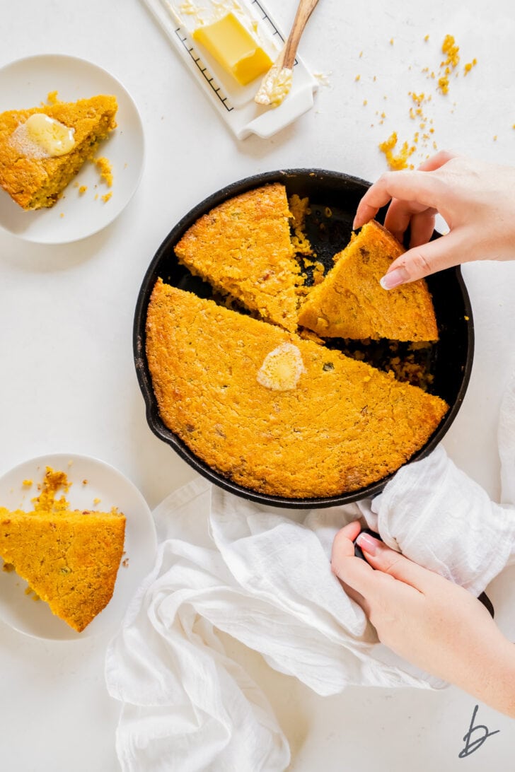 hand picking up slice of jalapeno cheese cornbread from a skillet with two plates with cornbread next to skillet