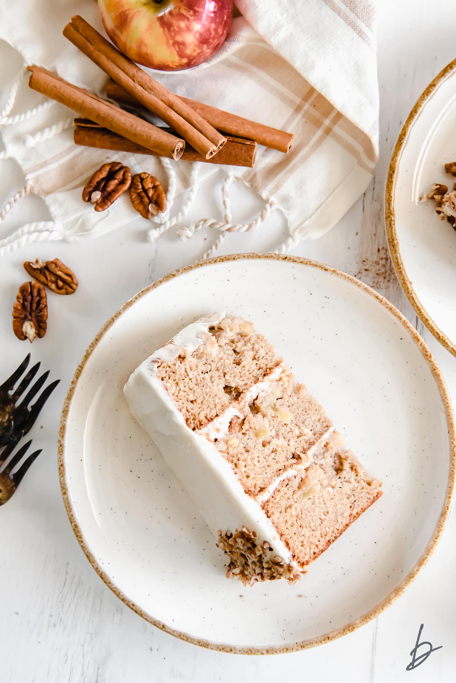 slice of triple layer apple spice cake on white gold-rimmed plate next to cinnamon sticks and pecans