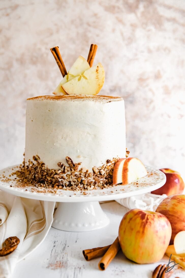 layer apple spice cake with cream cheese frosting garnished with chopped pecans, cinnamon sticks and apple slices on a white cake plate