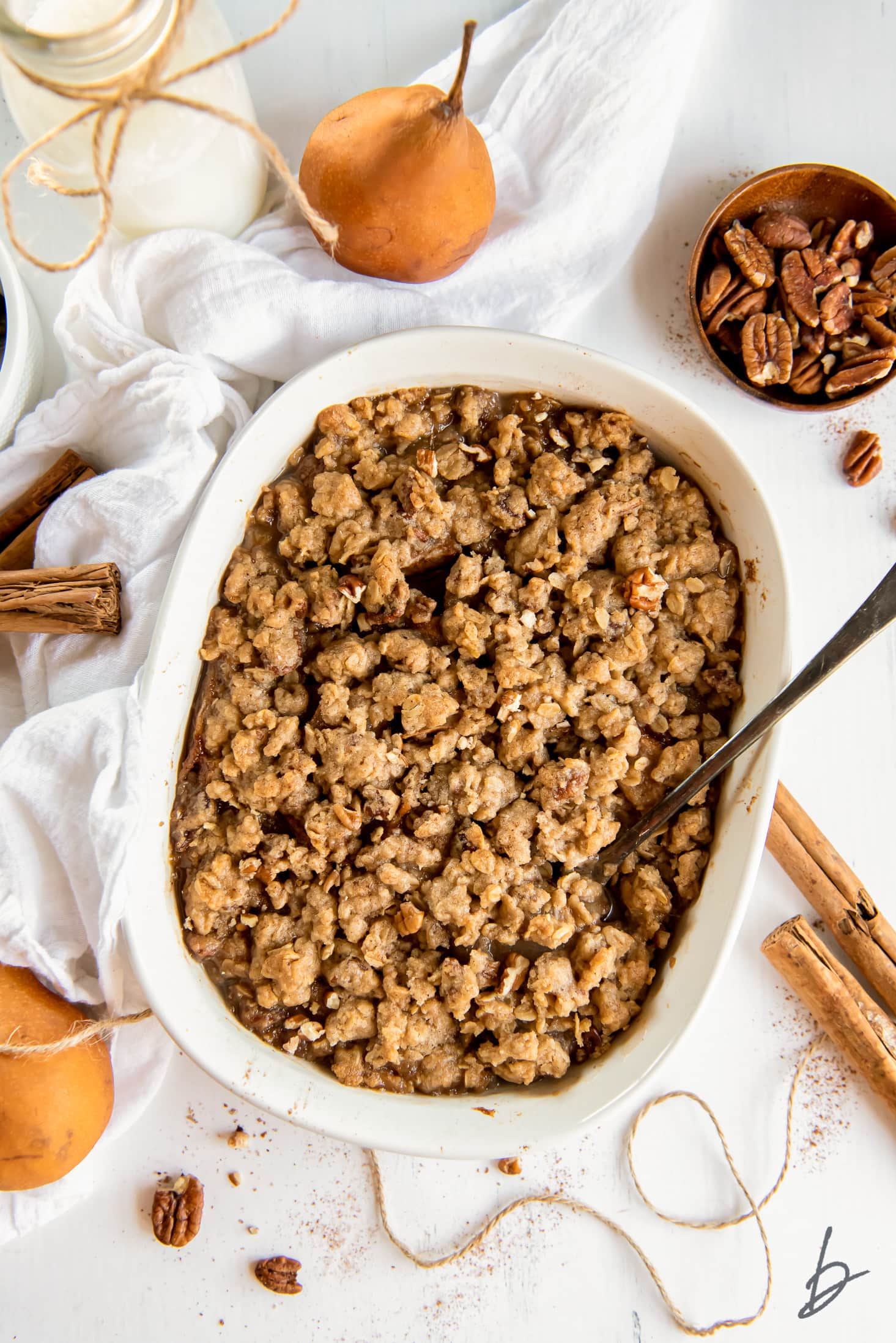 Pear crisp in a baking dish next to white cloth, pears and cinnamon sticks.