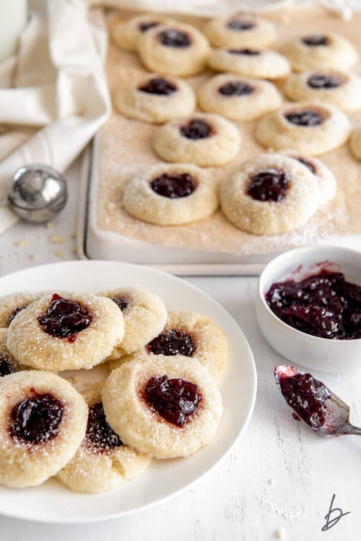 plate of jam thumbprint cookies next to bowl of raspberry jam in front of cookie sheet with more cookies