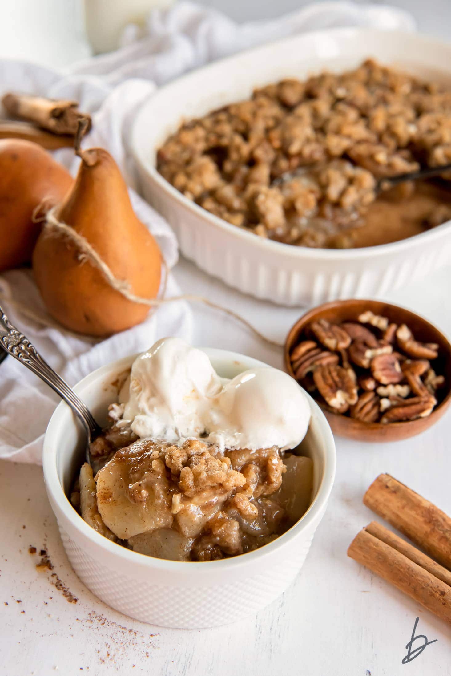 pear crisp in a bowl with vanilla ice cream next to pears, pecans and baking dish.