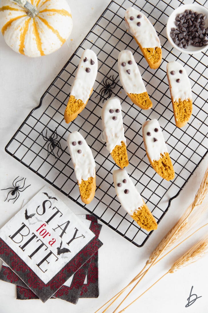 white chocolate dipped pumpkin biscotti that look like ghosts on black wire cooling rack next to halloween napkin and mini pumpkin